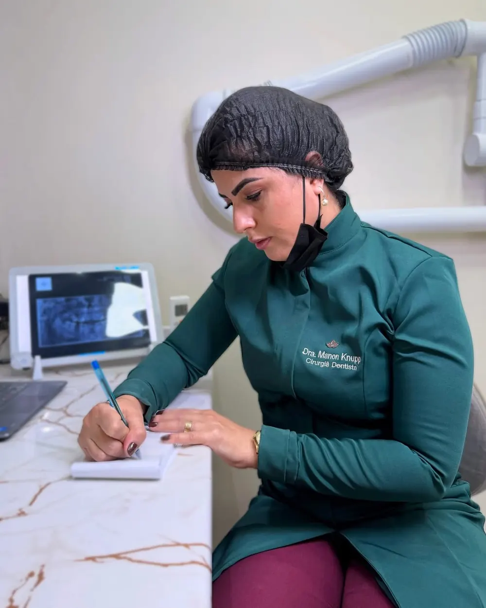 Dra. Manon Knupp fazendo anotações no consultório, com radiografia na tela ao fundo